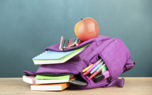 Purple backpack with school supplies on wooden table on green desk background